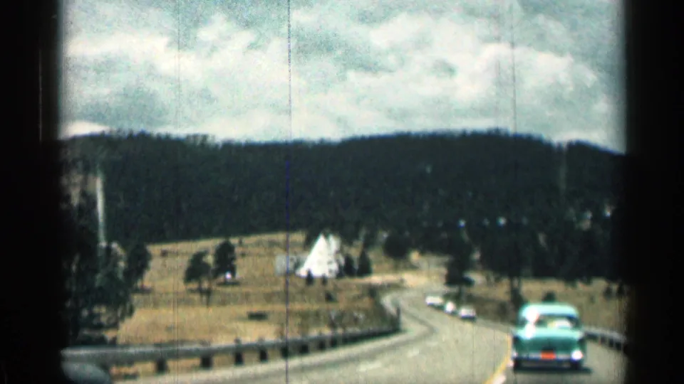 1963 Colorado home-movie frame from a moving car looking down a road through open meadow, with a white triangular roadside structure, guardrail, distant vehicles, wooded hills, and cloudy sky.