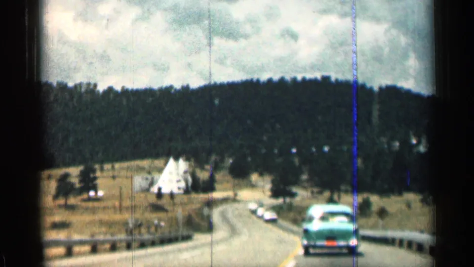 1963 Colorado 8mm frame showing the turquoise sedan ahead on a curving road through meadowland, with guardrails, distant vehicles, a white triangular structure, wooded hills, and a cloudy sky.