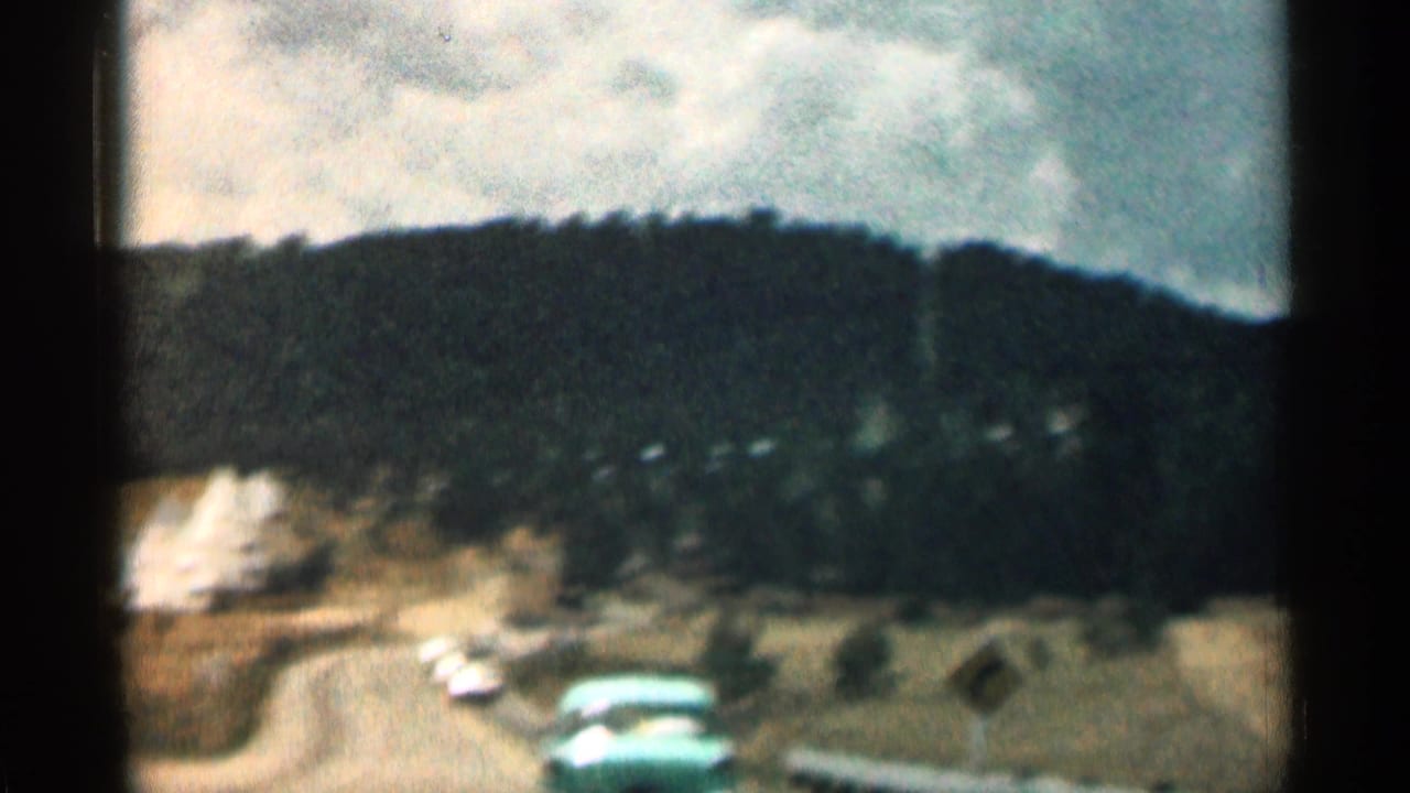 Winding road with cars below forested hills, seen from inside a moving vehicle.