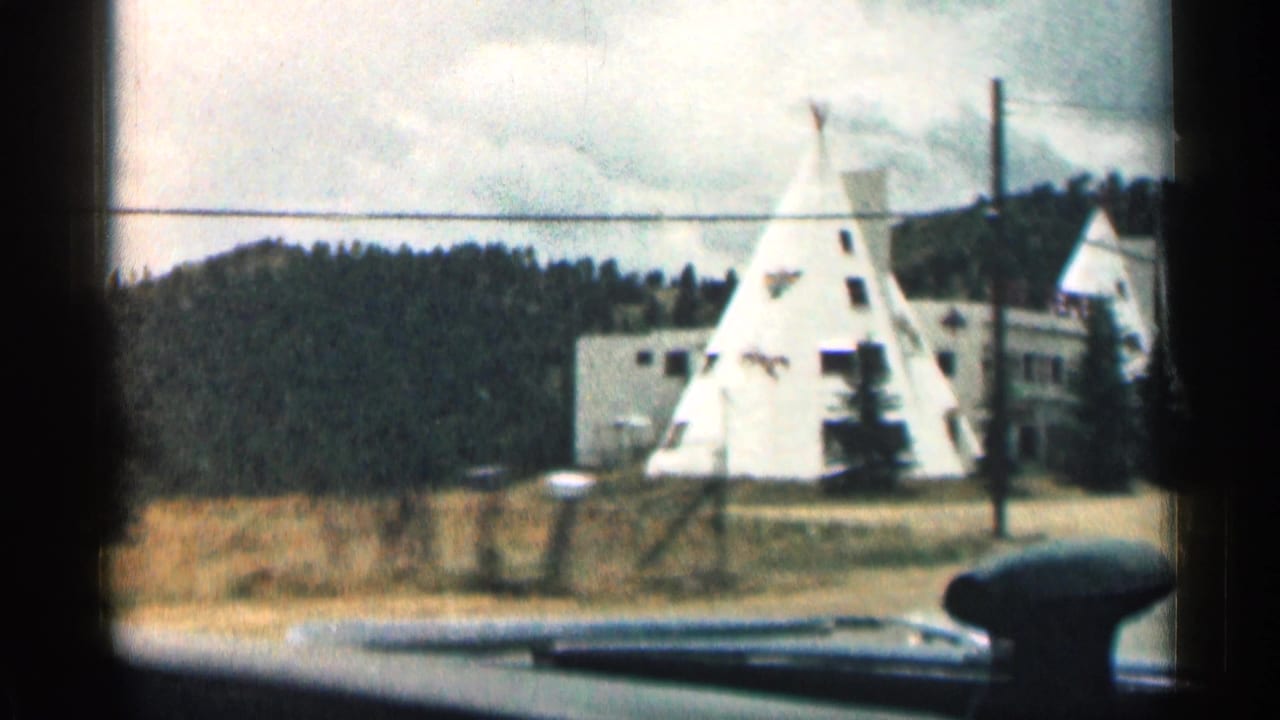White teepee-shaped roadside building beside trees, seen past a car dashboard.
