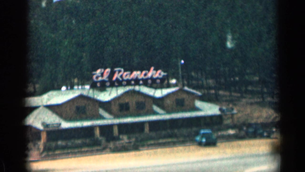 Mid-century El Rancho roadside restaurant in Colorado, with neon-style sign, parked cars, highway edge, and wooded hillside.