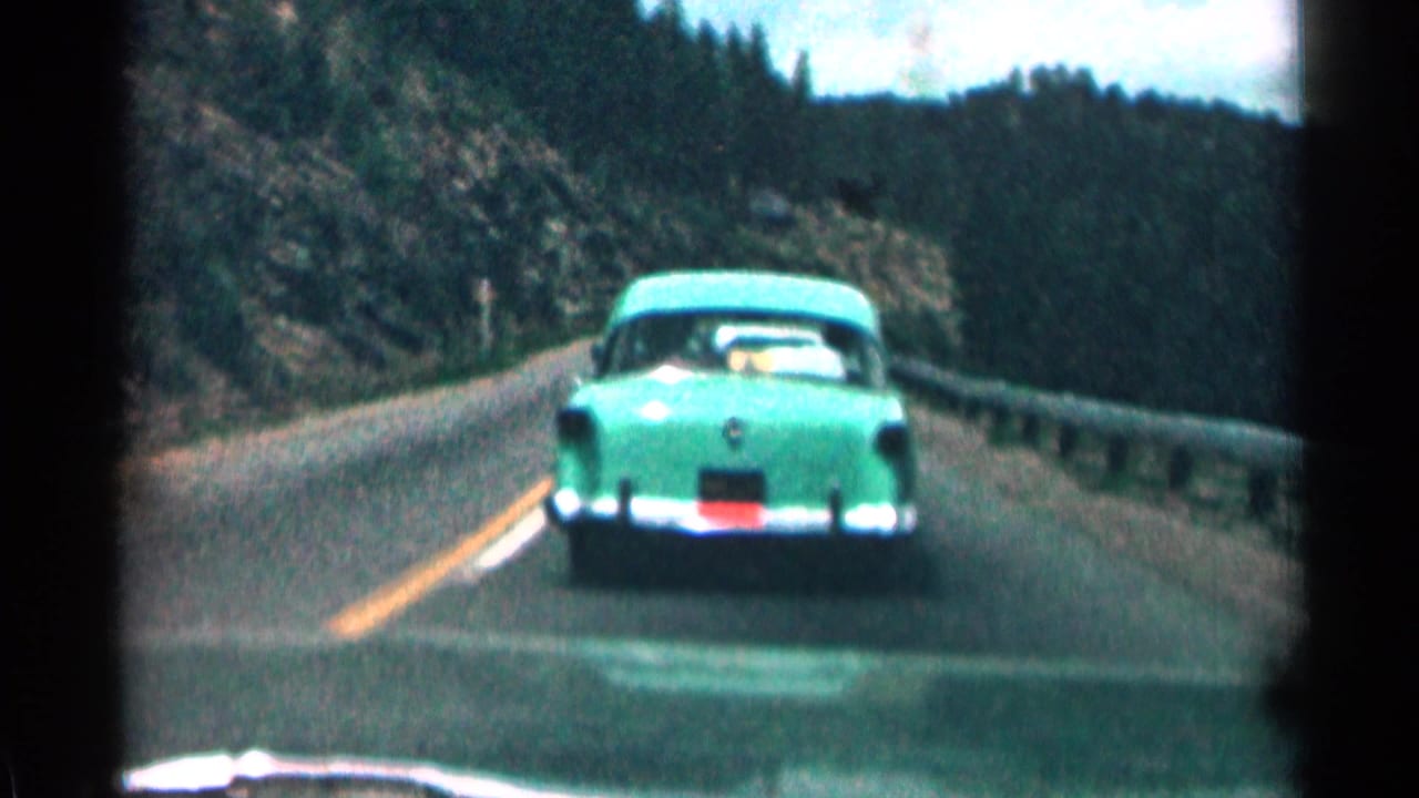 Green 1950s sedan seen from behind on a winding Colorado mountain highway in 1963 8mm home-movie footage.