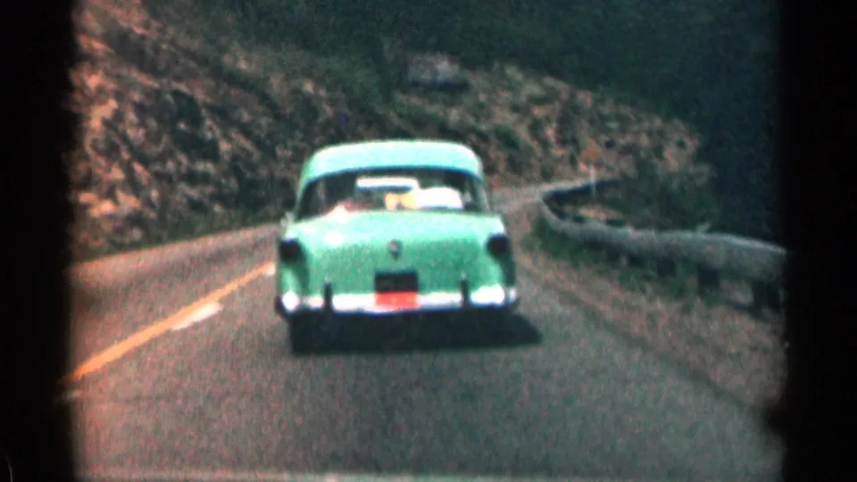 Green 1950s sedan approaches a bend on a Colorado mountain highway in grainy color 8mm footage.