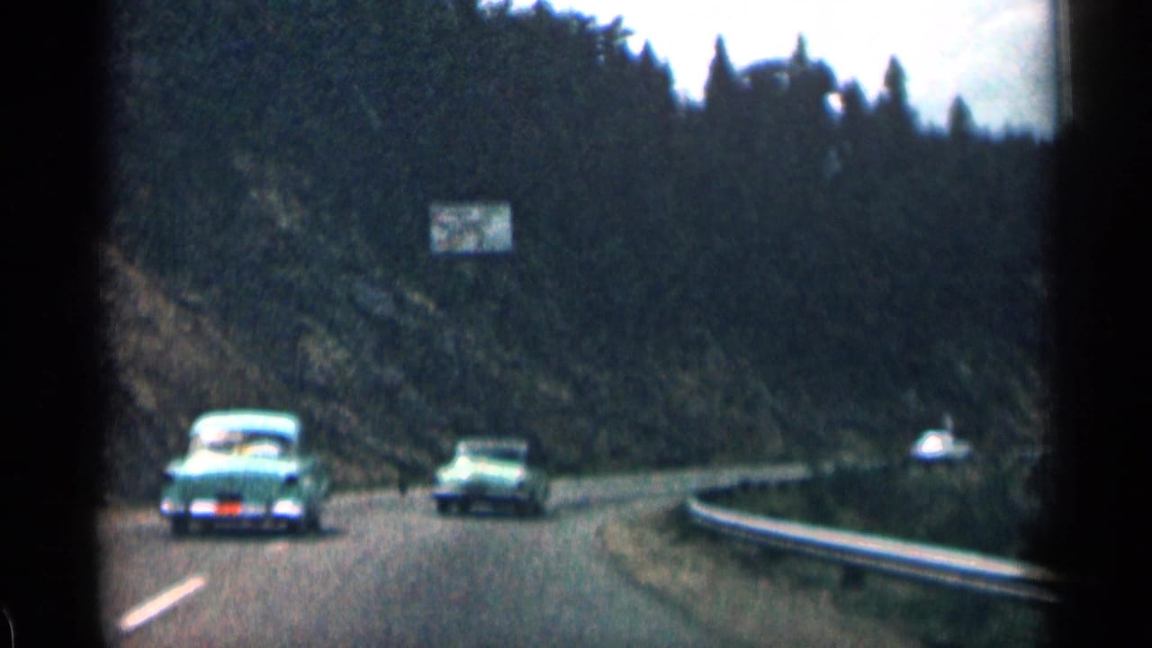Classic cars drive along a curving mountain road bordered by forest and guardrail.