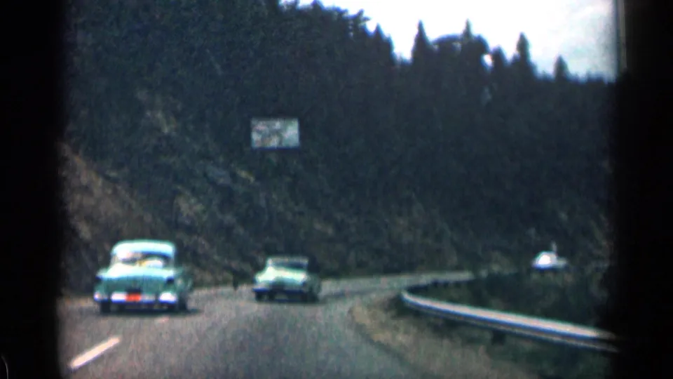 1963 Colorado road scene with classic cars on a winding mountain route, evergreen trees, a guardrail, and a small roadside sign.