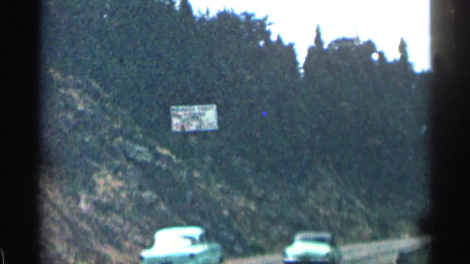 1963 Colorado 8mm frame showing cars below a roadside sign on a curving road cut into a wooded mountain slope.
