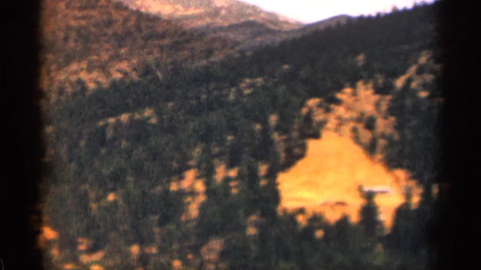1963 Colorado home-movie frame of forested hills and a bright clearing seen from an elevated roadside viewpoint.