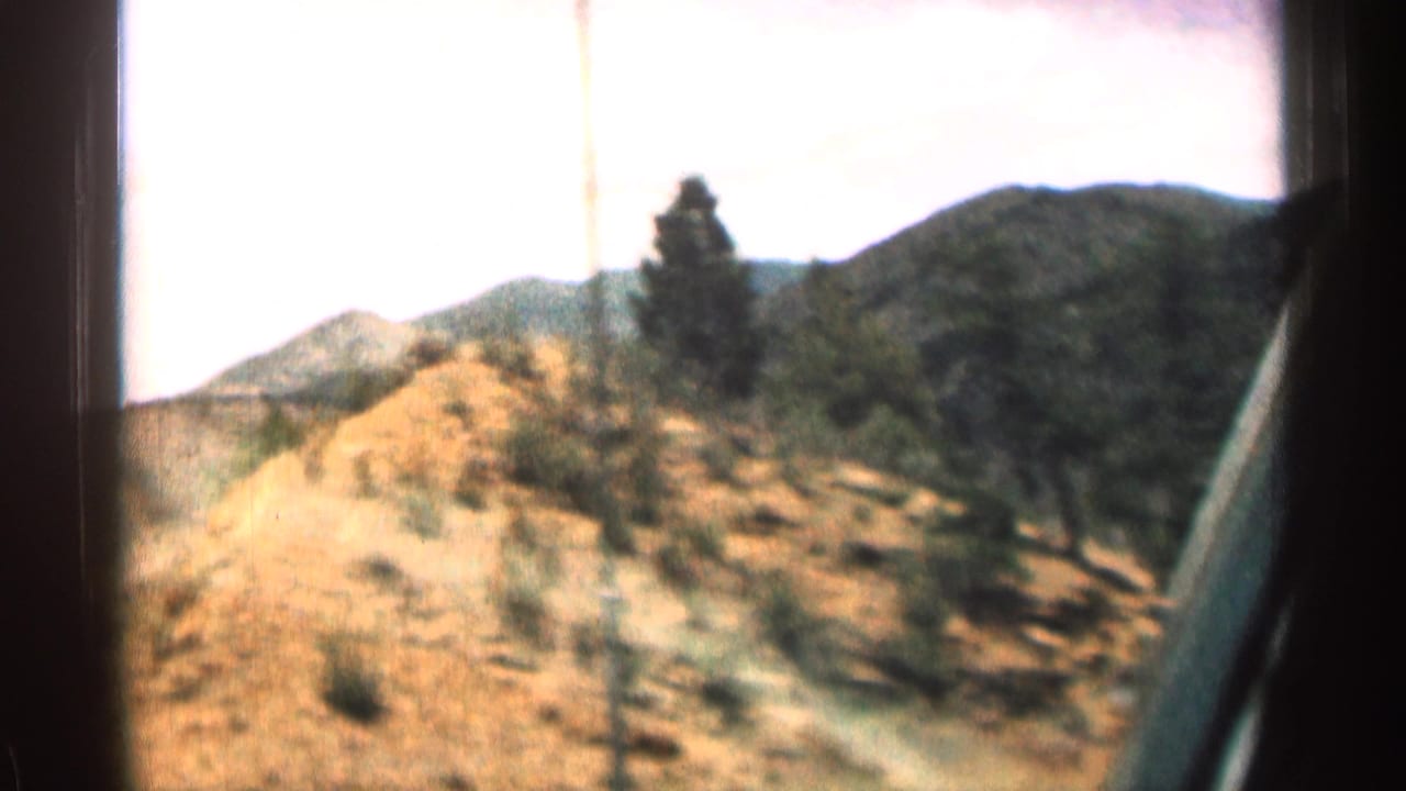 1963 Colorado home-movie view from inside a vehicle, looking past window framing toward dry hills and forested mountain terrain.
