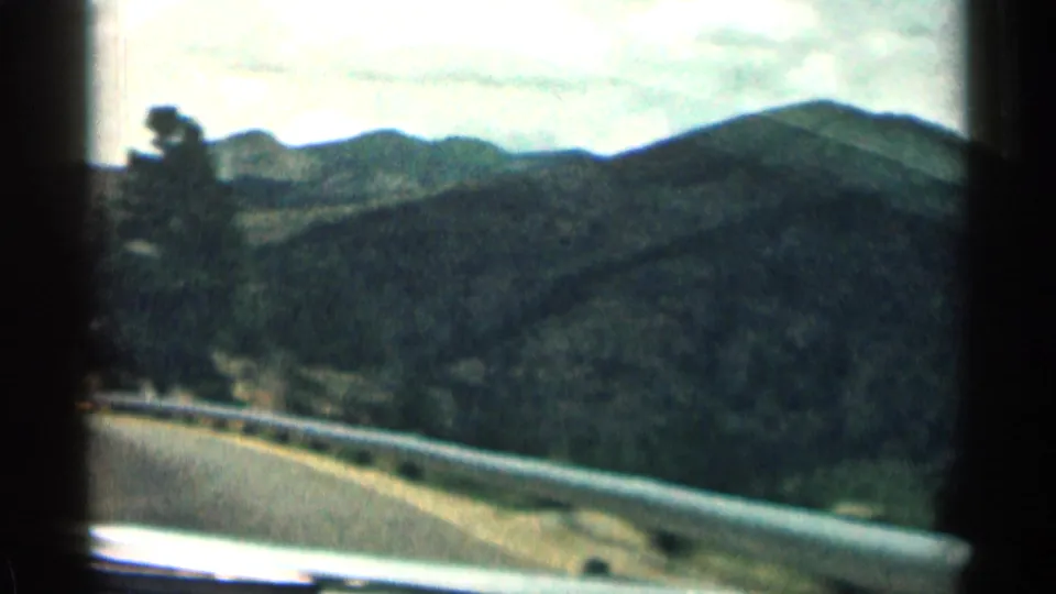 1963 Colorado road-trip footage shows a curving paved road, metal guardrail, and wooded mountain slopes under a cloudy sky.