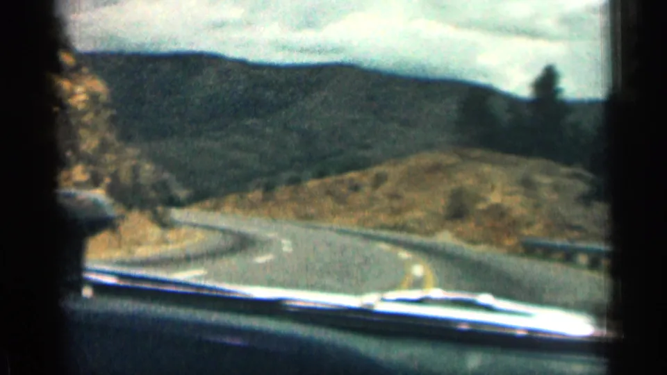 The vehicle approaches a bend on a Colorado mountain road, with dry slopes, guardrails, distant wooded hills, and windshield framing visible.