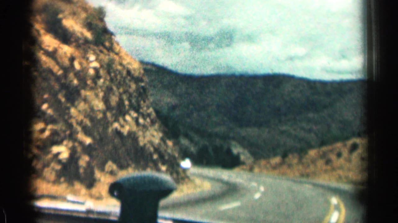 Winding mountain road seen through a car windshield beside rocky cliffs and distant hills.