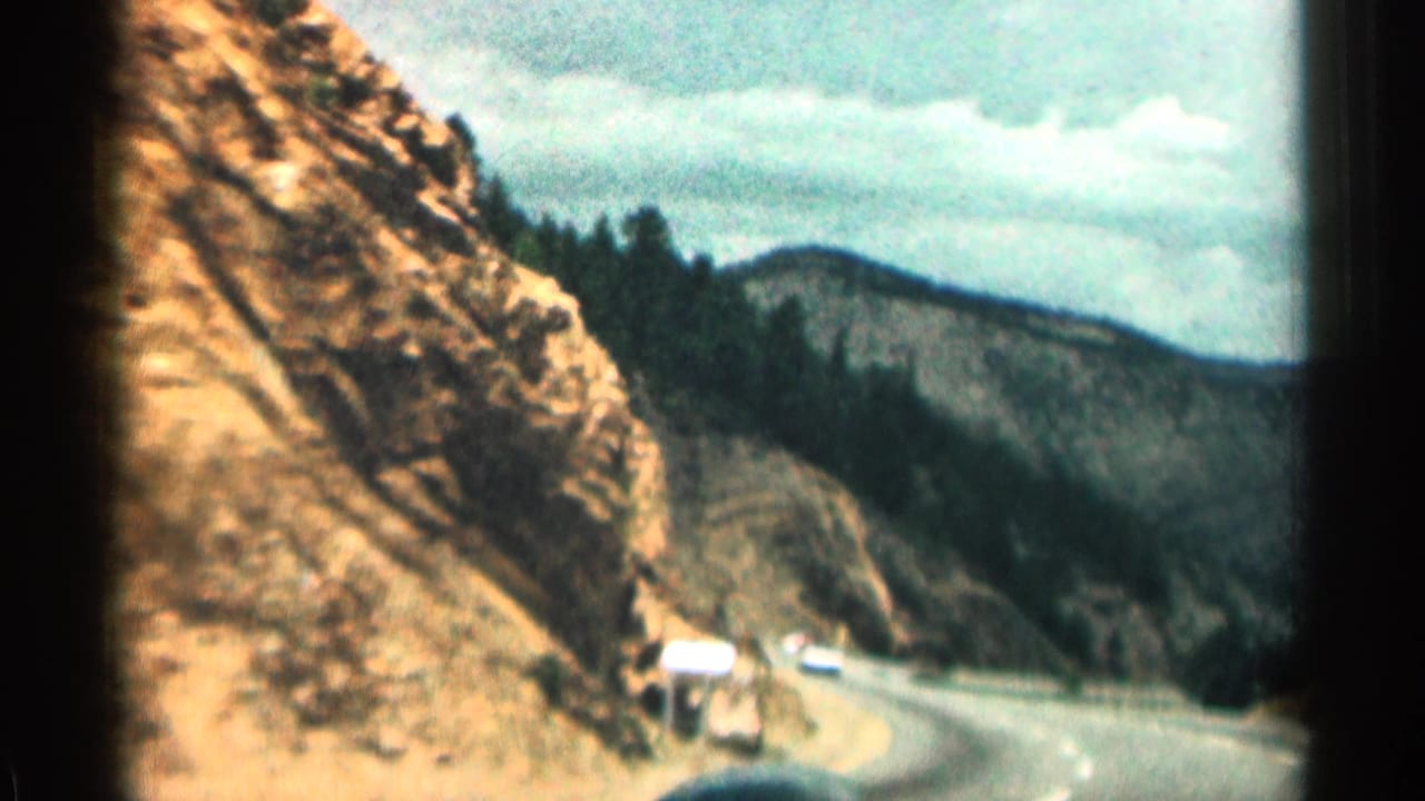 View from a moving car along a Colorado mountain road, with a steep rock wall, distant forested hills, cloudy sky, and small vehicles ahead.