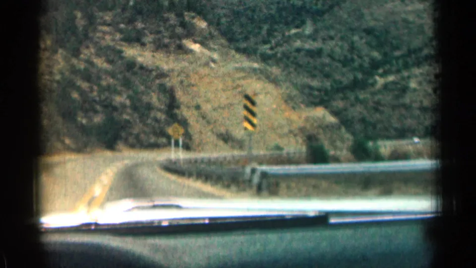 1963 Colorado car-window view approaching a mountain road curve with a guardrail, road sign, striped marker, rocky slope, and dashboard edge.