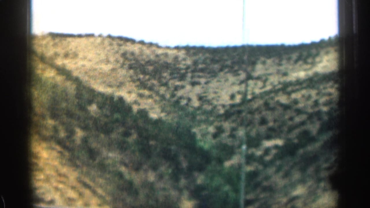 1963 Colorado 8mm frame showing a scrub-covered rocky hillside and bright sky from a moving vehicle.