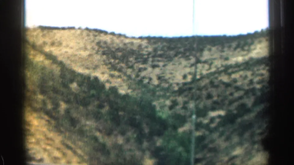 1963 Colorado 8mm frame showing a scrub-covered rocky hillside and bright sky from a moving vehicle.