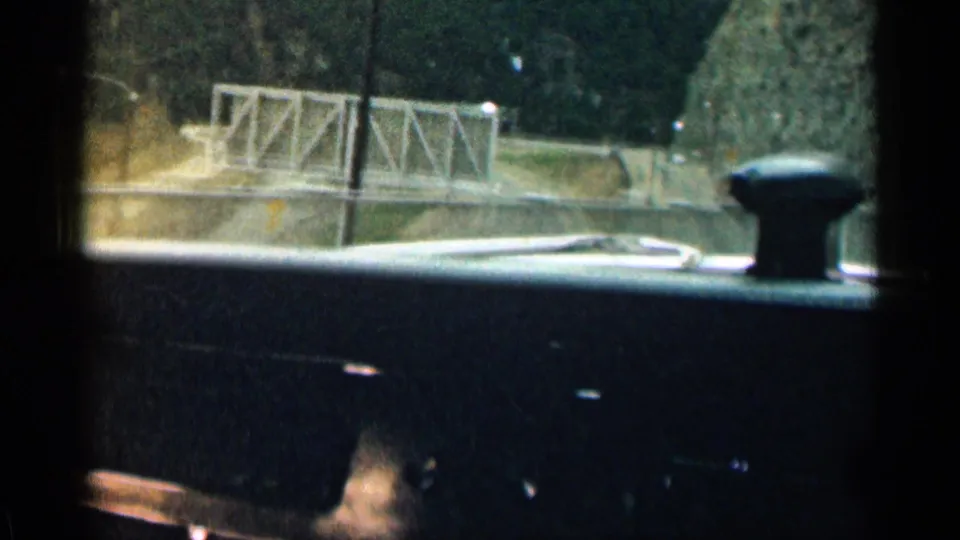 1963 Colorado 8mm frame showing a mountain road, metal guardrail or small bridge, hillside, and the edge of the car interior.