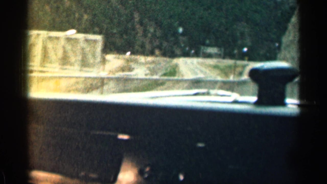 1963 Colorado 8mm frame from inside a vehicle, showing a road edge, guard wall, and dark evergreen slope beyond.