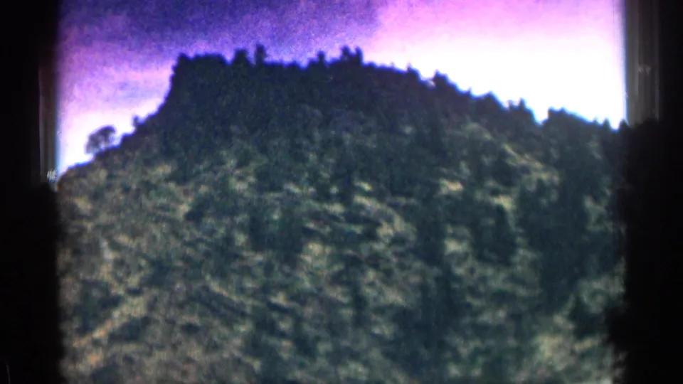 1963 Colorado mountain scenery with a tree-lined ridge and rocky hillside captured from a moving car.