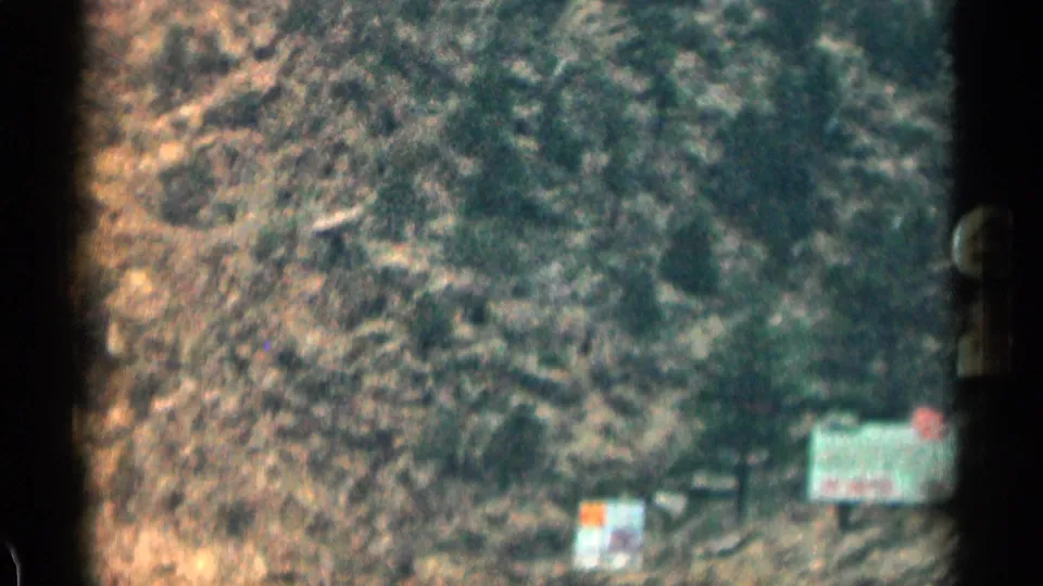 Colorado mountain road scene with a rocky cliff face, scattered trees, and small roadside signs at the base.