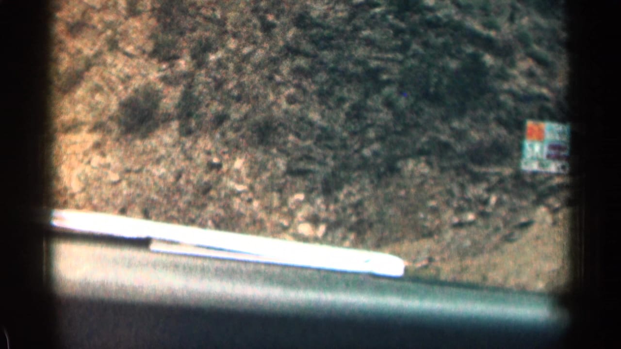 1963 Colorado 8mm road-trip frame from inside a car, looking past the hood toward a rocky roadside curve and a small sign ahead.