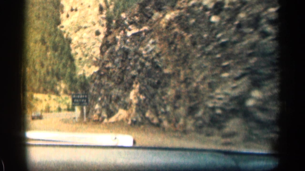 Road through rocky canyon with a roadside sign and car hood visible.