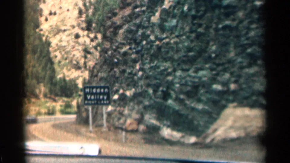 1963 Colorado road-trip frame showing a Hidden Valley sign beside a curving road, with rocky canyon walls and trees beyond the car hood.