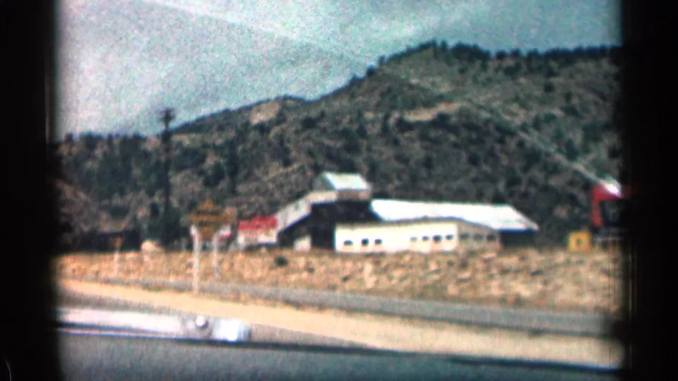 Colorado road-trip frame from inside a car showing a roadside building or structure, open ground, signs, and wooded hills in the background.