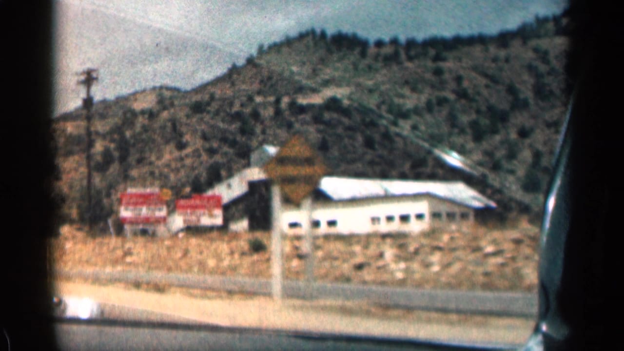 1963 8mm Colorado road-trip frame showing a low white roadside building, red signs, a utility pole, and scrubby mountain slopes through a car window.