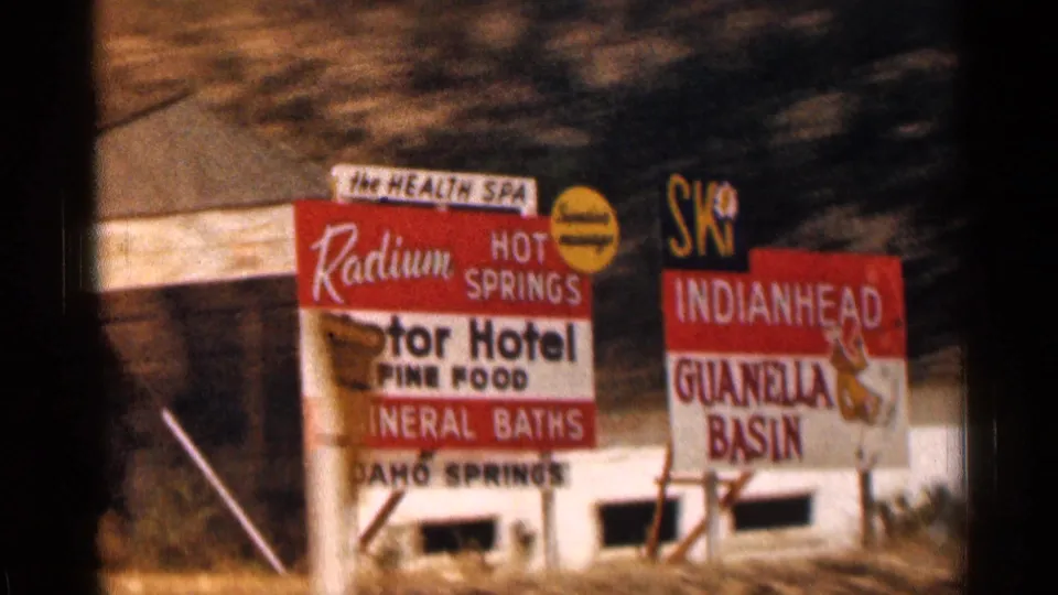 1963 Colorado road-trip frame with prominent roadside signs reading Radium Hot Springs, hotel, mineral baths, Idaho Springs, and Guanella Basin.