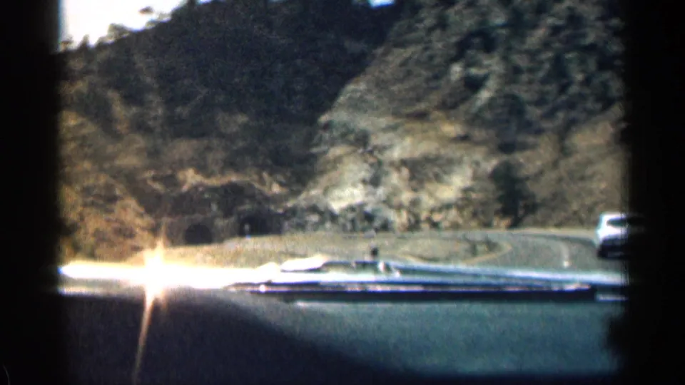 1963 8mm Colorado driving view over the car dashboard toward a curving mountain road, rocky slopes, and a vehicle at the edge of the frame.