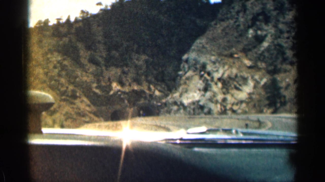View through a car windshield on a winding mountain road with rocky slopes ahead.