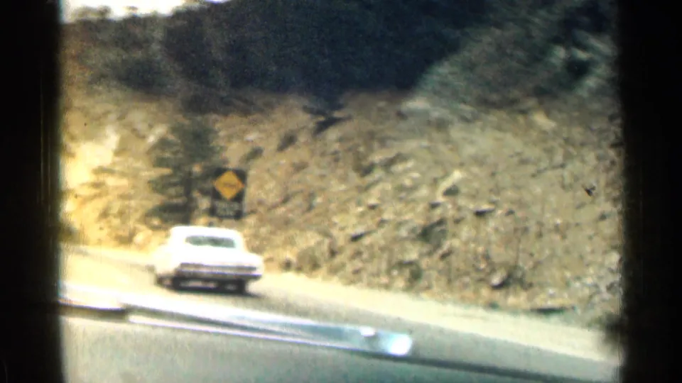 A white car drives ahead on a Colorado mountain road near a yellow tunnel warning sign in 1963 home-movie footage.