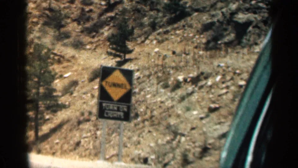 Close roadside view of a yellow tunnel warning sign against a rocky Colorado hillside, filmed from inside a moving car.