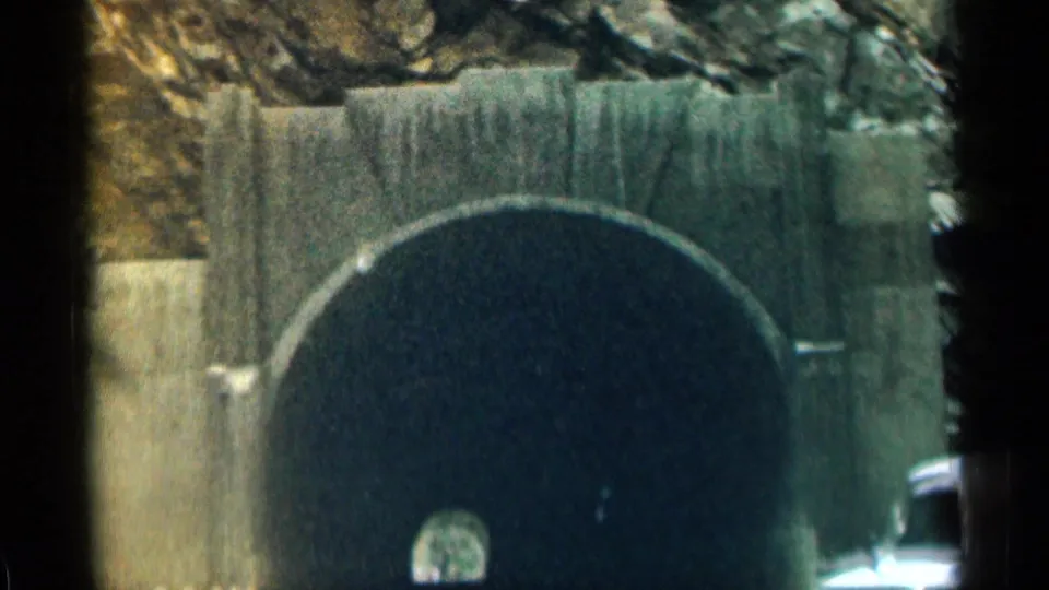 The car approaches a dark stone tunnel entrance on a Colorado mountain road in grainy 1963 8mm footage.