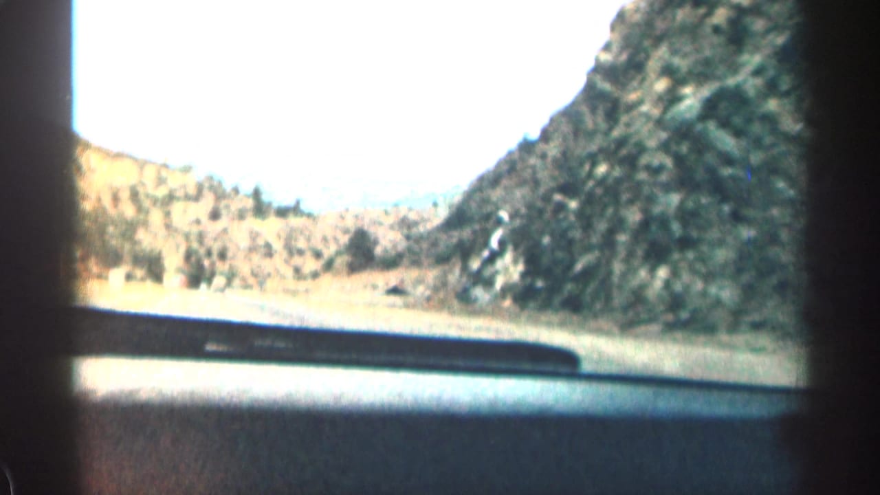 1963 Colorado home-movie frame from inside a car, looking past the dashboard toward a curving road and rugged hills.