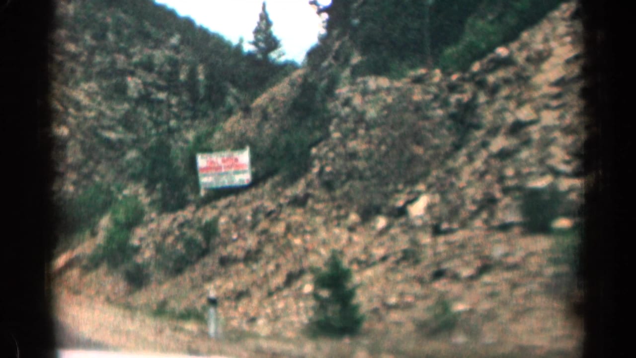 1963 Colorado 8mm frame showing a canyon road from inside a car, with rocky slopes, trees, and a small roadside sign ahead.