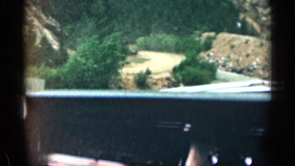 1963 Colorado home-movie view from inside a car, looking over the dashboard toward a pale road, shrubs, and rocky canyon terrain.