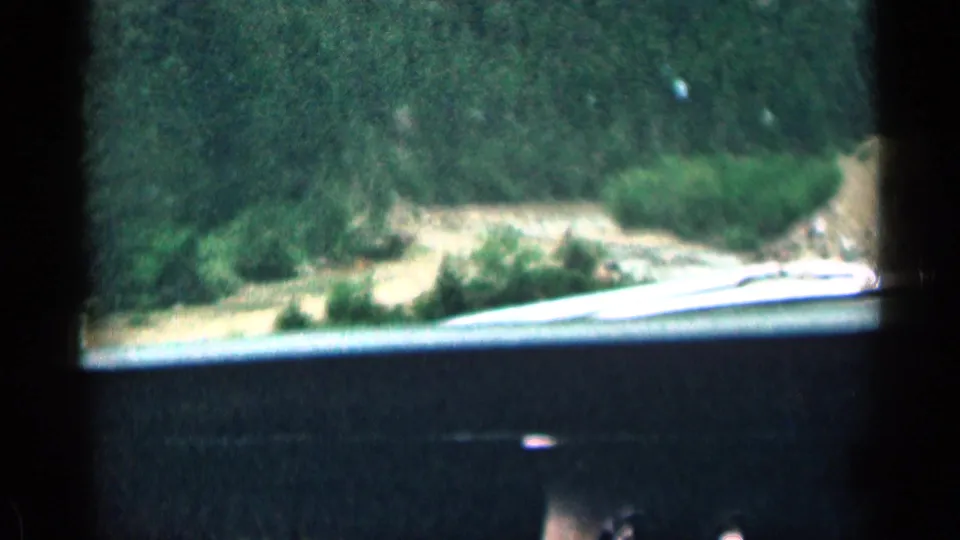 1963 Colorado 8mm frame showing a moving-car view across the dashboard toward shrubs, a pale road, and forested canyon walls.