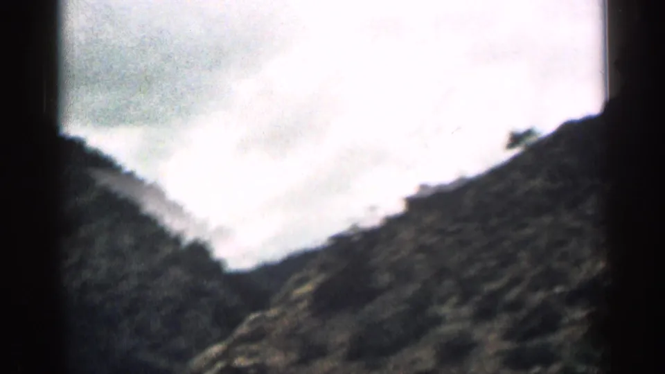 1963 Colorado home-movie frame with overexposed sky above dark, rugged mountain slopes during an in-car drive.