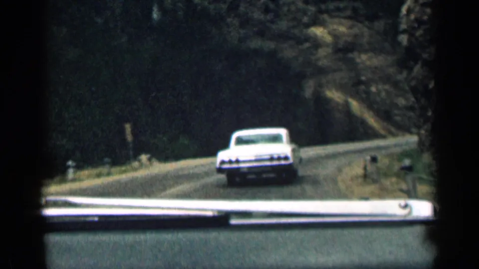 1963 Colorado 8mm frame from inside a car following a white period automobile on a curving mountain road.