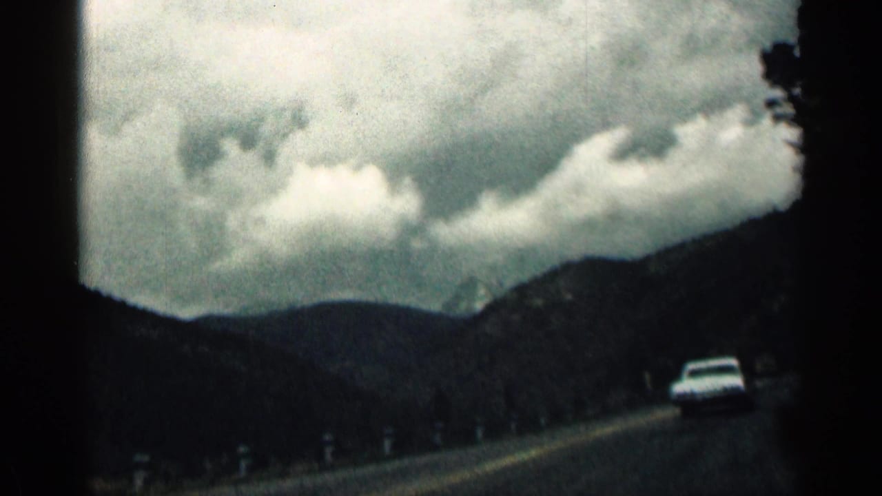 1963 Colorado 8mm frame from inside a moving car, showing a white car ahead on a paved mountain road under heavy clouds.