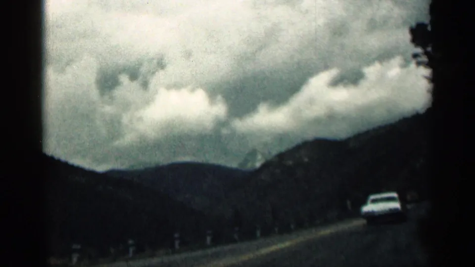 1963 Colorado 8mm frame from inside a moving car, showing a white car ahead on a paved mountain road under heavy clouds.