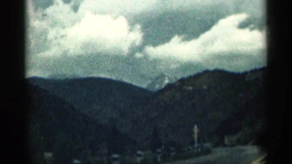 1963 Colorado 8mm frame with cloudy sky, layered mountain ridges, and a road edge visible from a passenger-view driving shot.