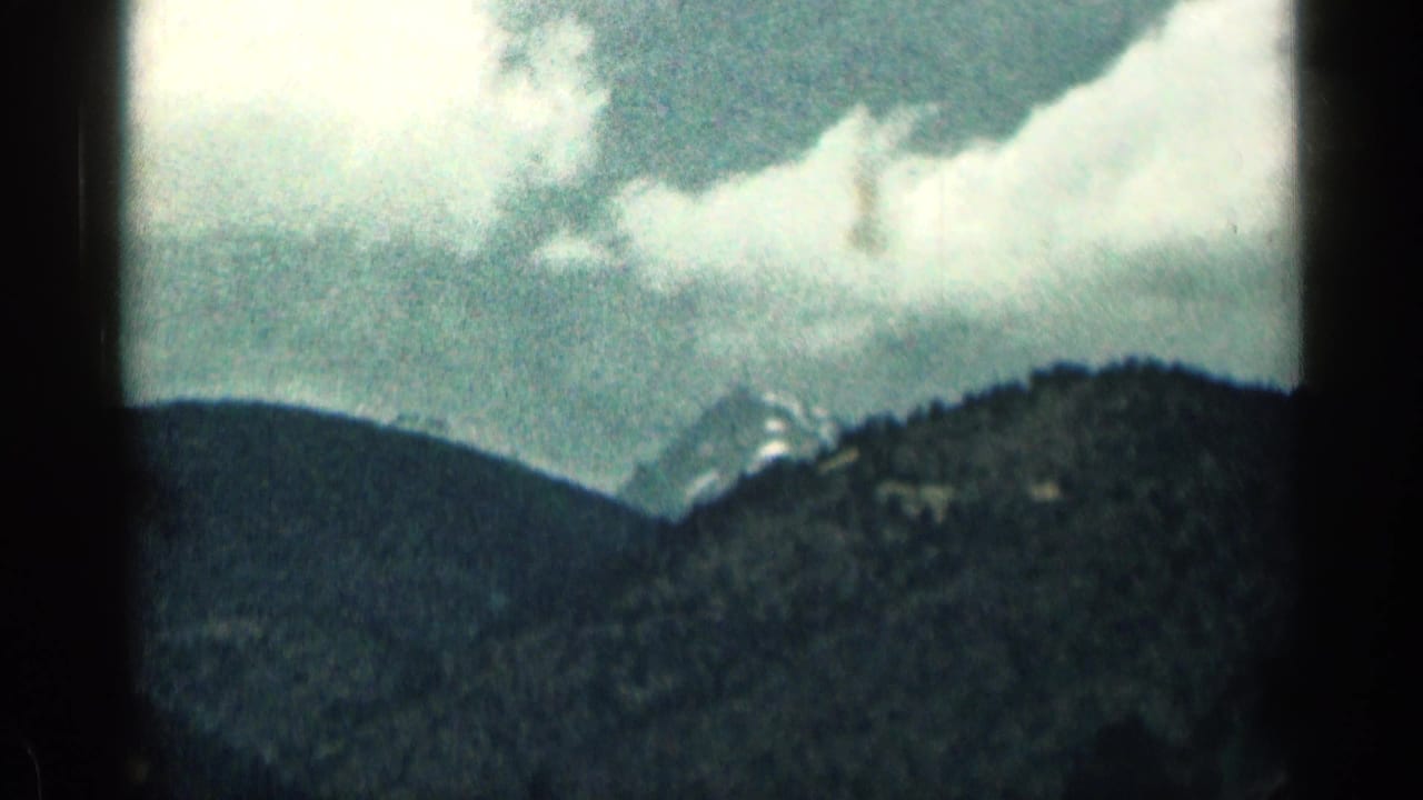 1963 Colorado 8mm frame showing cloudy sky above forested mountains with distant snowy peaks softened by haze.