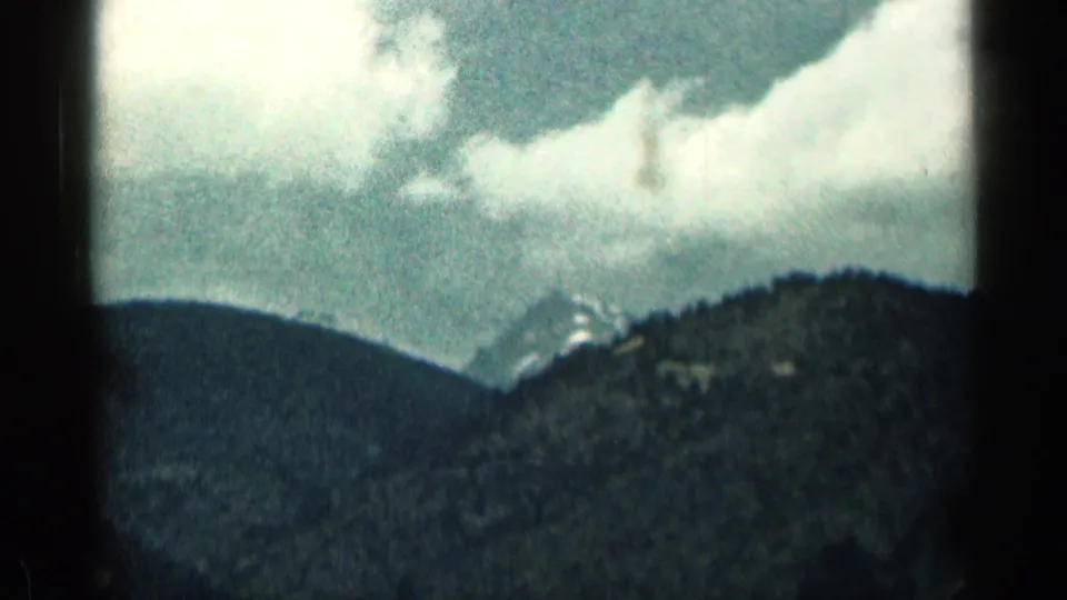 1963 Colorado 8mm frame showing cloudy sky above forested mountains with distant snowy peaks softened by haze.