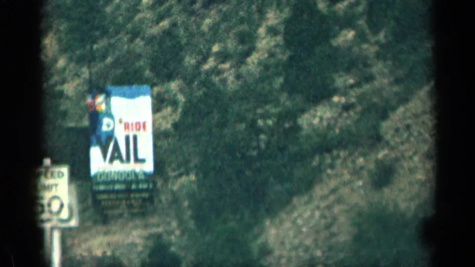 1963 Colorado roadside frame showing a Ride Vail gondola billboard beside rocky terrain and a partial speed sign.