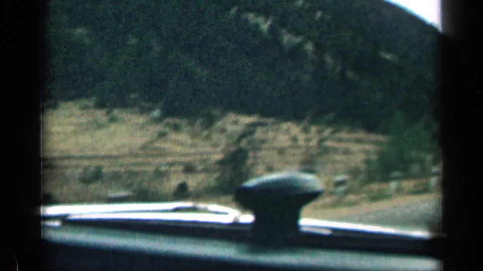 Handheld in-car road view in Colorado, with dashboard foreground, riverbank, roadside curve, and shadowed trees on the hillside.