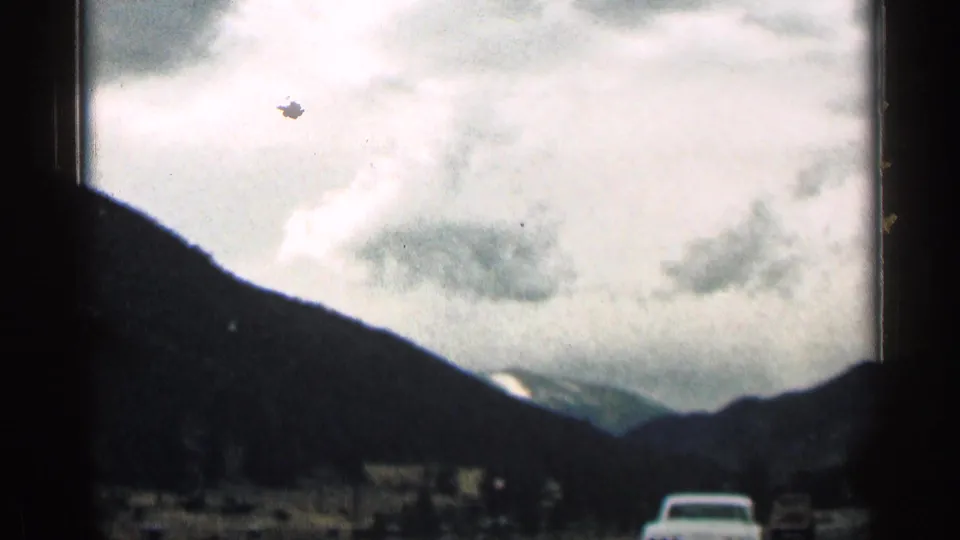 1963 Colorado road-trip frame showing cars ahead on a mountain road, dark slopes, open sky, and cloudy weather from inside a vehicle.
