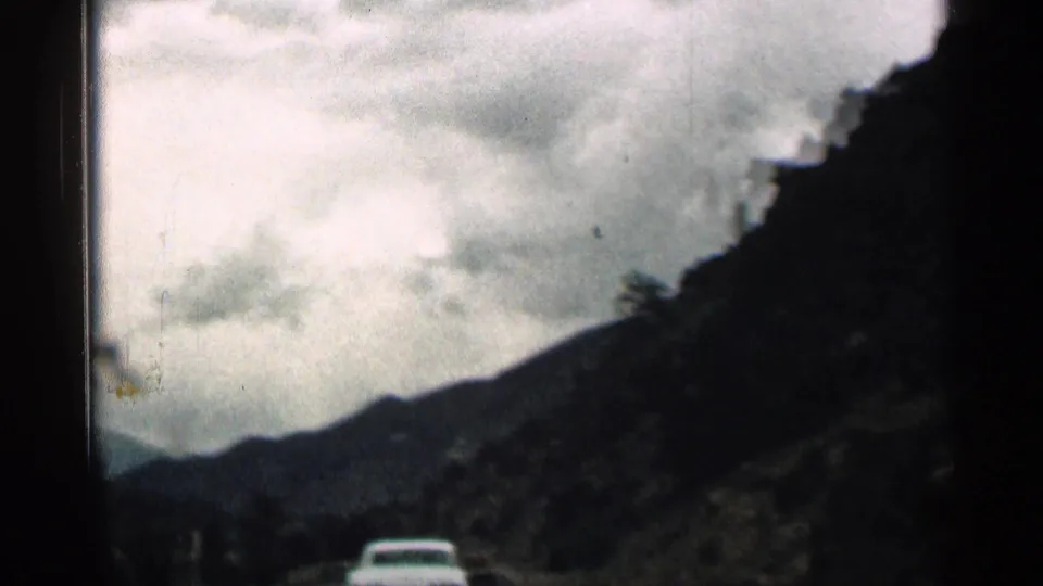 Passenger-view 8mm frame from Colorado, with a white car ahead between dark mountain slopes and low clouds.