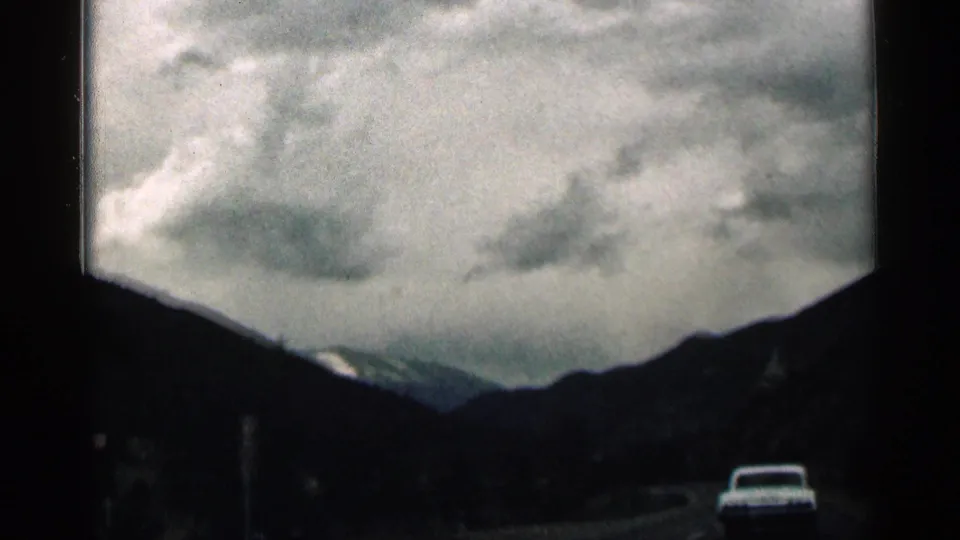 Vintage 8mm Colorado road-trip frame showing a car on a mountain road beneath layered storm clouds.